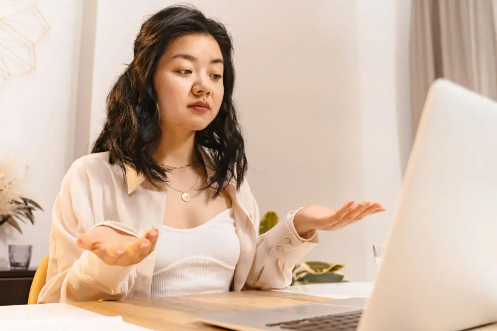 Jeune femme perplexe devant son ordinateur, les mains ouvertes en signe de question